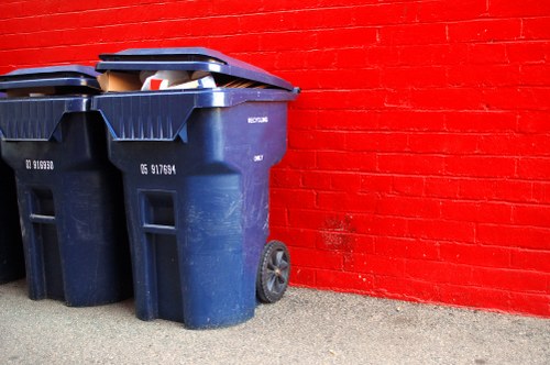 Two large dark blue plastic wheelie bins with black lids positioned on a grey concrete pavement next to a bright red brick wall. The bins are intended for rubbish collection and are placed outdoors, with one bin slightly in front of the other and their lids partly open, revealing some waste materials inside. The bins have visible wheels at the back for mobility and are situated in a residential or urban area. The scene appears well-lit, with natural daylight illuminating the textured surface of the brick wall and the smooth surface of the bins. This image exemplifies typical rubbish collection equipment used by House Clearance Norwood for waste disposal services in the Norwood area, showcasing the practical aspect of household rubbish storage before removal or disposal.