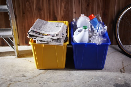 Two large plastic bins placed on a concrete floor against a wooden wall, with the yellow bin on the left filled with stacked newspapers and magazines, and the blue bin on the right containing clear plastic bottles, empty bottles, a white plastic container, and other plastic packaging. The scene appears to be in a garage or storage area, with part of a bicycle wheel visible on the right side. The containers are used for rubbish collection, aligned with household waste removal services provided by House Clearance Norwood, serving the Norwood area. The lighting is neutral, highlighting the textures of the plastic and paper, emphasizing the materials commonly associated with household refuse disposal and recycling activities in the local area.