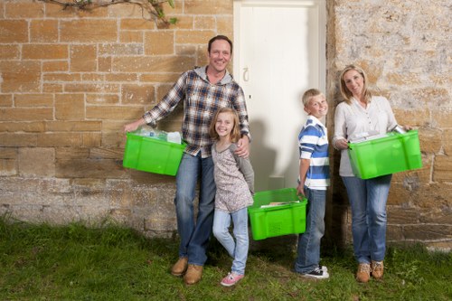 A family of four standing outside a stone and brick wall, each holding a bright green plastic storage box. The father is positioned on the left, smiling, with the box held in his right hand, wearing a checked shirt and jeans. Next to him is a young girl with curly hair, wearing a blue and white patterned sweater and jeans, smiling and holding a box in front of her. To her right are a boy in a blue and white striped shirt and jeans, standing with a neutral expression, holding a box with both hands, and a woman on the far right, dressed in a white blouse and jeans, also smiling and holding a green storage box. The scene appears to be in an outdoor residential area, possibly on a driveway or garden space, with grass on the ground and the family preparing for rubbish collection or clearance, consistent with services offered by House Clearance Norwood in the local area near Norwood and the surrounding postcode region.