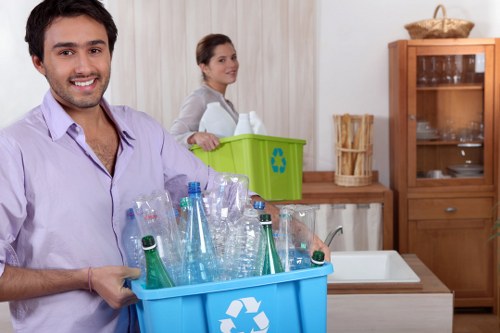 In a domestic kitchen setting, a smiling young man in a light purple shirt is holding a blue recycling bin filled with various clear plastic bottles, some toppled over, ready for disposal. Behind him, a woman dressed in white is near a green recycling container, which features a blue recycling symbol, and appears to be preparing or organizing waste materials. The kitchen has a wooden cabinet with glass doors and a small wicker basket on top, positioned against a light-colored wall with vertical paneling. The workspace includes a white sink with a chrome tap and a beige countertop, with a wooden dish rack visible on the side. The environment appears well-lit with natural daylight, emphasizing the clean, orderly appearance of the space, aligned with services offered by House Clearance Norwood in the area around Norwood, south London, where professional rubbish removal is needed for households or property clearances.